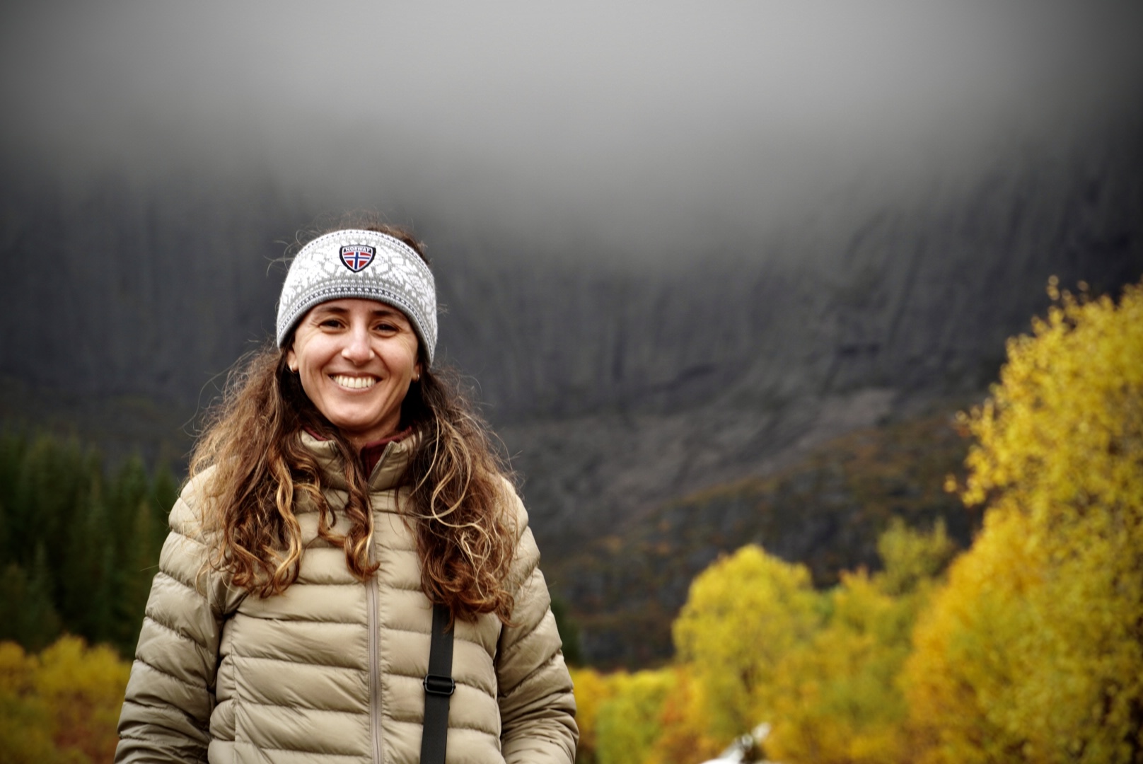 Smiling woman in a knit headband before yellow autumn trees and dark mountains.