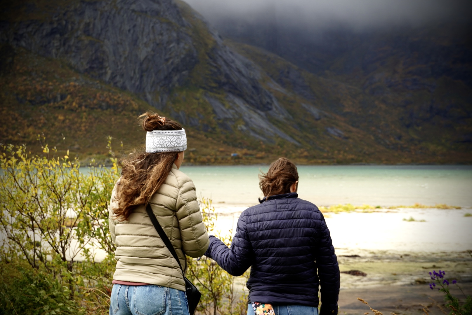 Two people walking beside pale coastal water below dark mountains.