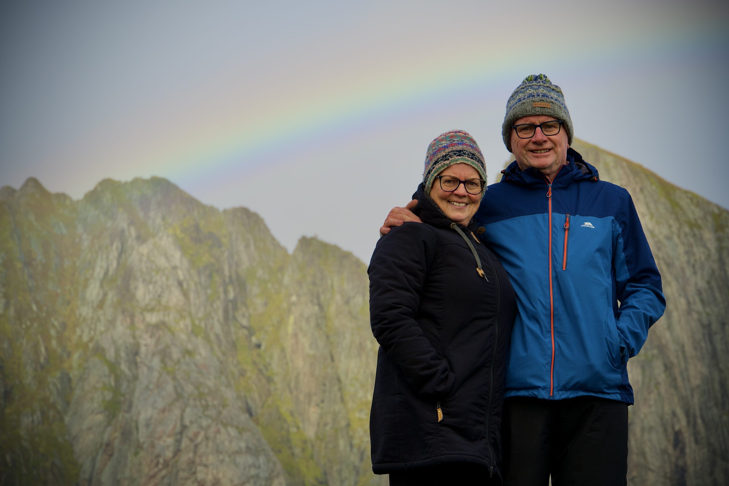 Two people posing in front of mountains beneath a faint rainbow.