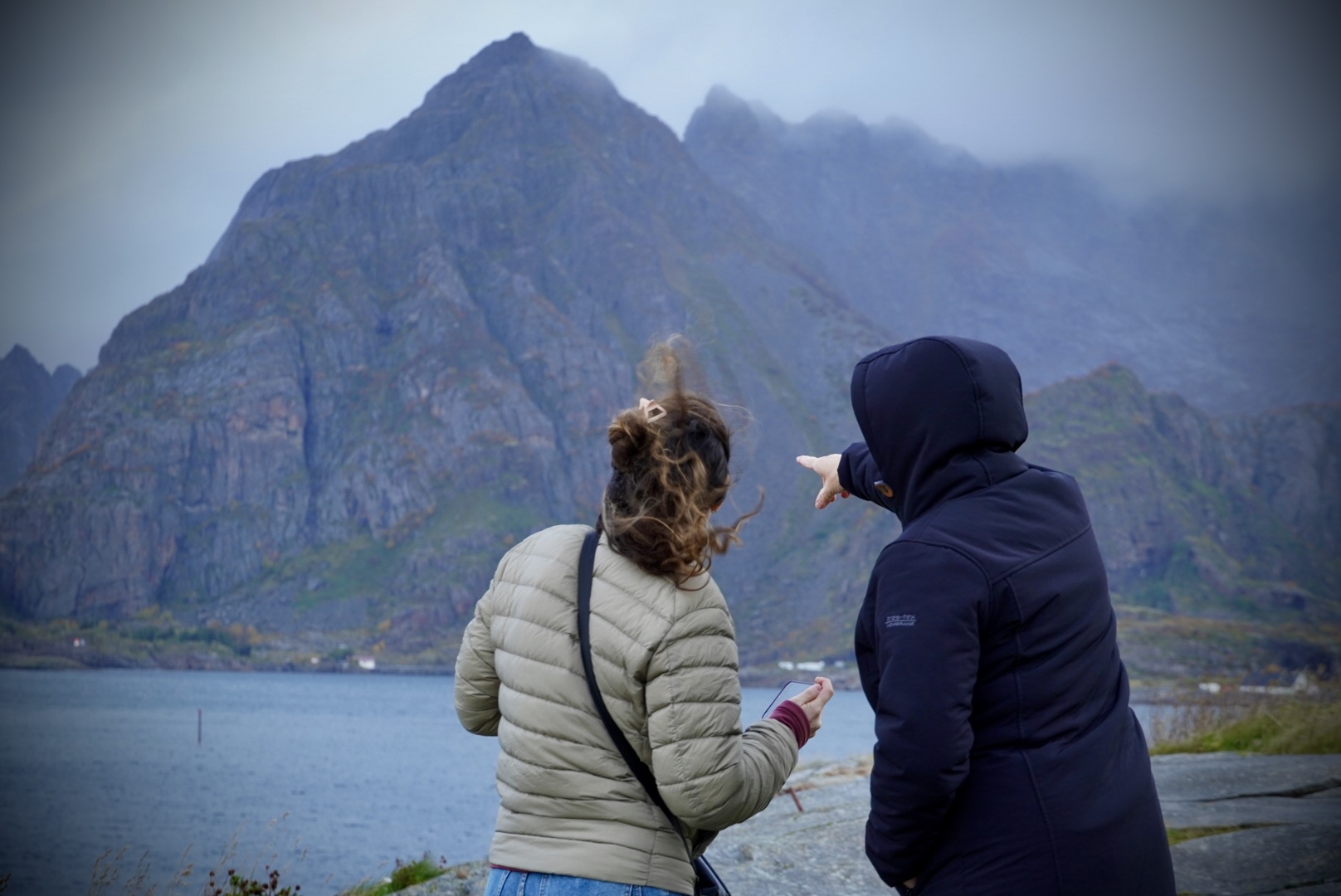 Two people in jackets looking toward mountain peaks across the water.