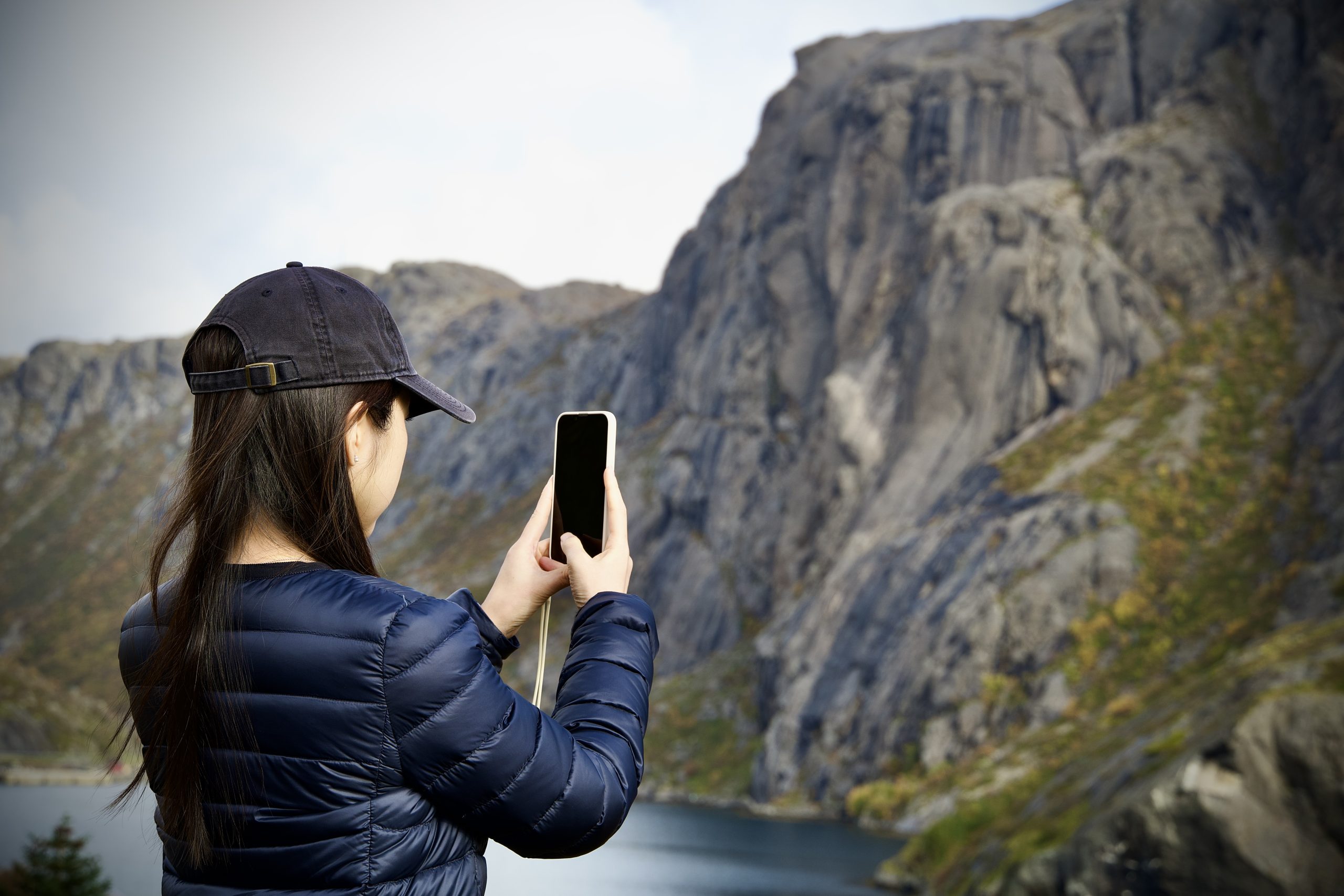 Person in a cap photographing a sheer rocky mountain by the water.